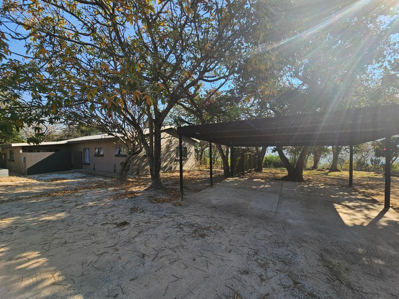 Exterior view of the property featuring a large carport, concrete driveway, and mature trees surrounding the building facade.