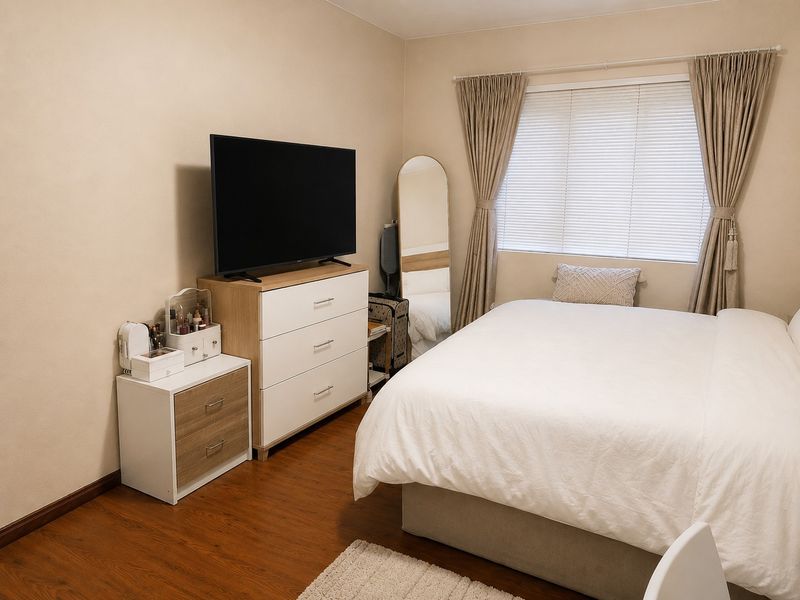A well-maintained bedroom featuring warm-toned laminate flooring and neutral wall finishes, complemented by functional window treatments including venetian blinds and curtains.