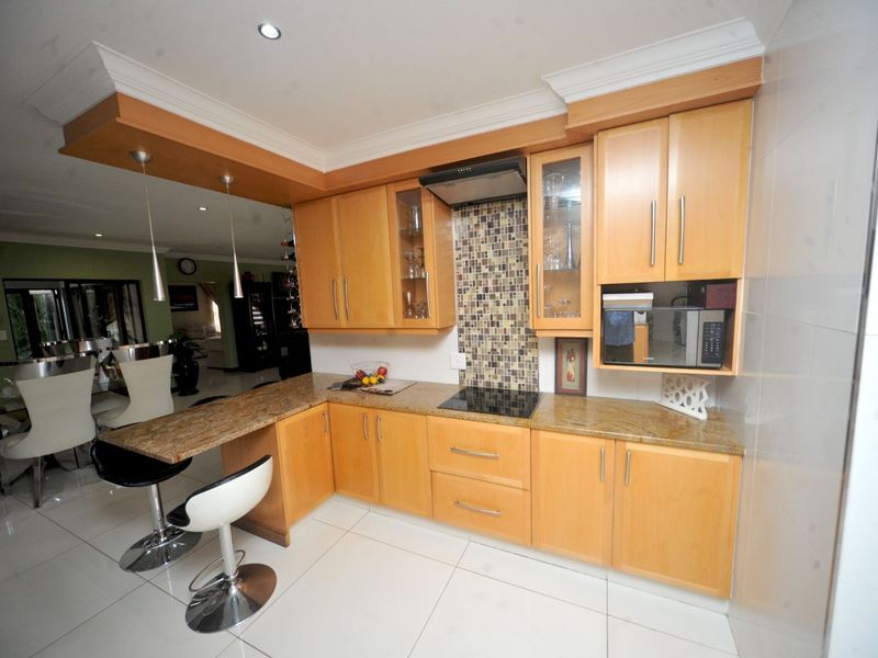 Modern kitchen featuring light wood cabinetry, granite countertops, mosaic tile backsplash, integrated appliances, and a breakfast bar overlooking the open-plan living area.