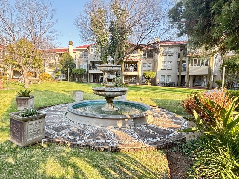 A well-maintained communal garden area featuring a central stone fountain, manicured lawns, and mature trees within a townhouse complex.