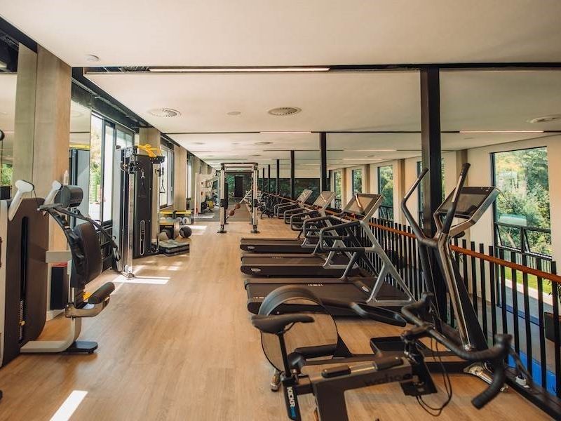 A modern, well-lit gym facility featuring mirrored walls, laminate flooring, and large windows overlooking the exterior, equipped with professional fitness machinery.