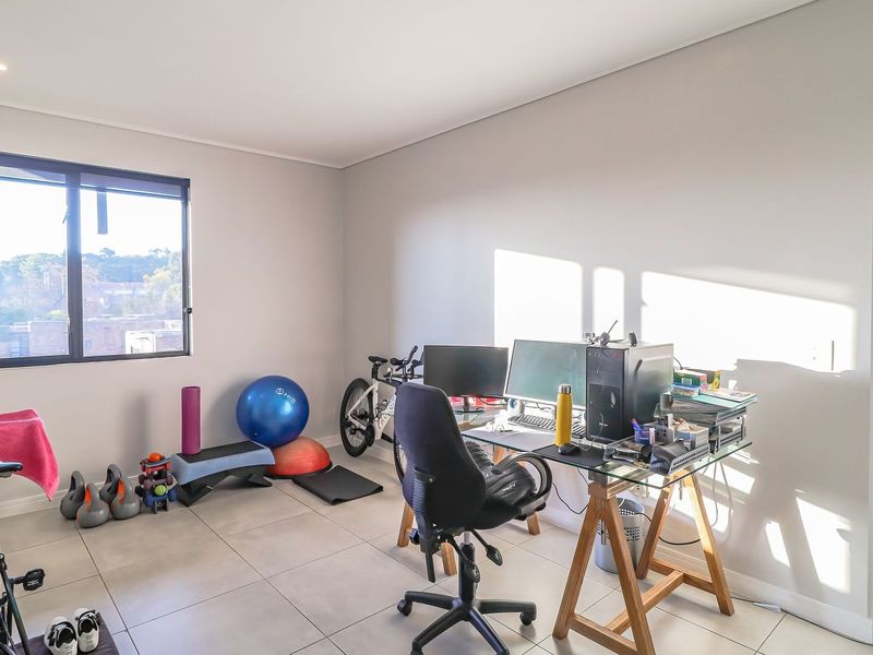 A bright, multi-purpose office space featuring neutral tiled flooring, recessed lighting, and large windows, currently utilized as a home workspace and gym area.