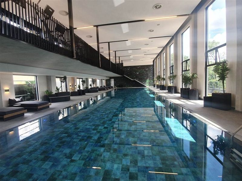 A modern indoor lap pool featuring large floor-to-ceiling windows, a mezzanine level, and high-quality tiled finishes in a sophisticated, well-maintained facility.