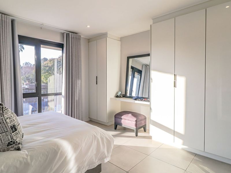 A bright, modern bedroom featuring extensive built-in cabinetry, neutral tiled flooring, and large windows providing natural light.