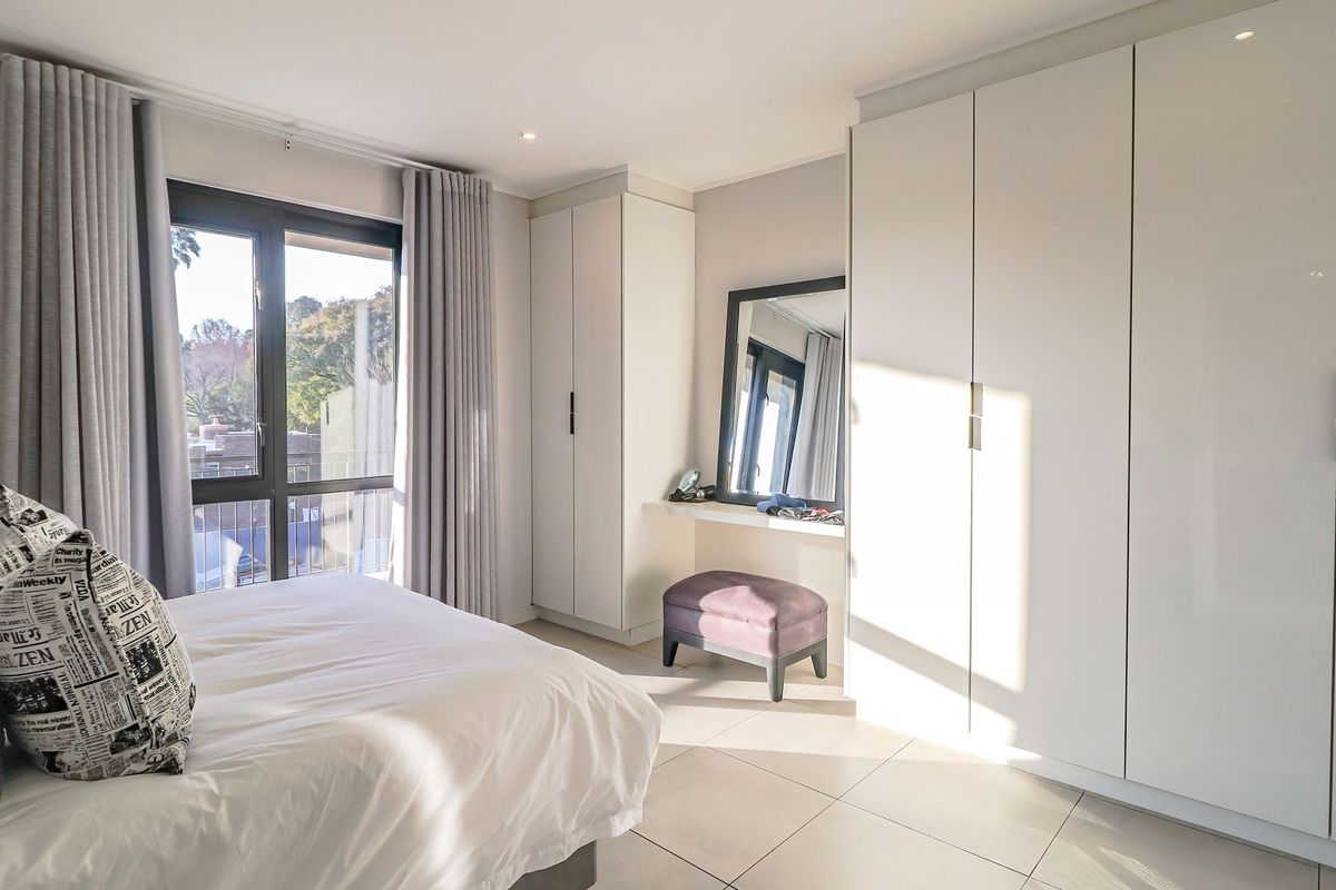 A bright, modern bedroom featuring extensive built-in cabinetry, neutral tiled flooring, and large windows providing natural light.