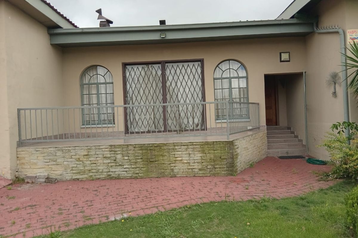 Rear exterior view featuring a raised patio with stone cladding, metal security gates, and arched windows, leading to a paved garden area.