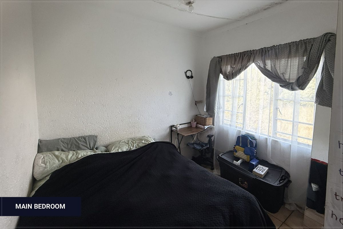 A bedroom featuring a window with curtains and tiled flooring, showing signs of ceiling water damage and wall wear.