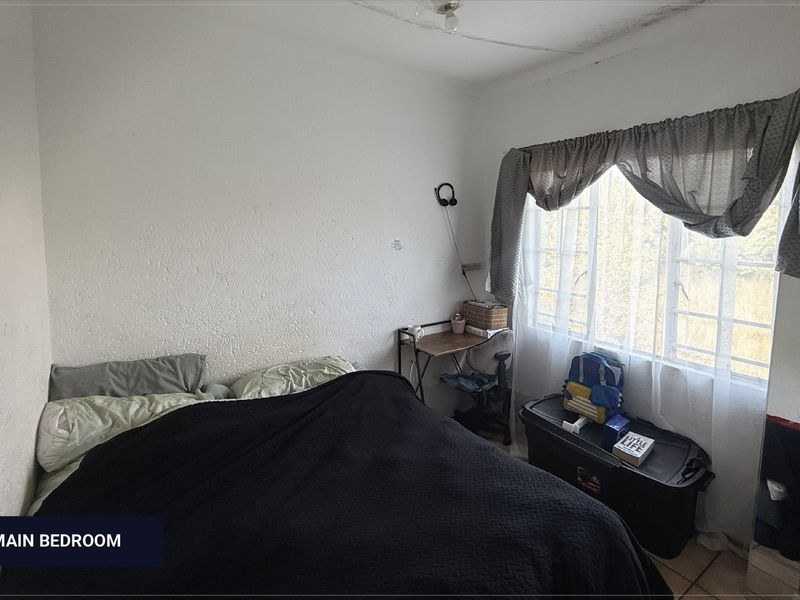 A bedroom featuring a window with curtains and tiled flooring, showing signs of ceiling water damage and wall wear.