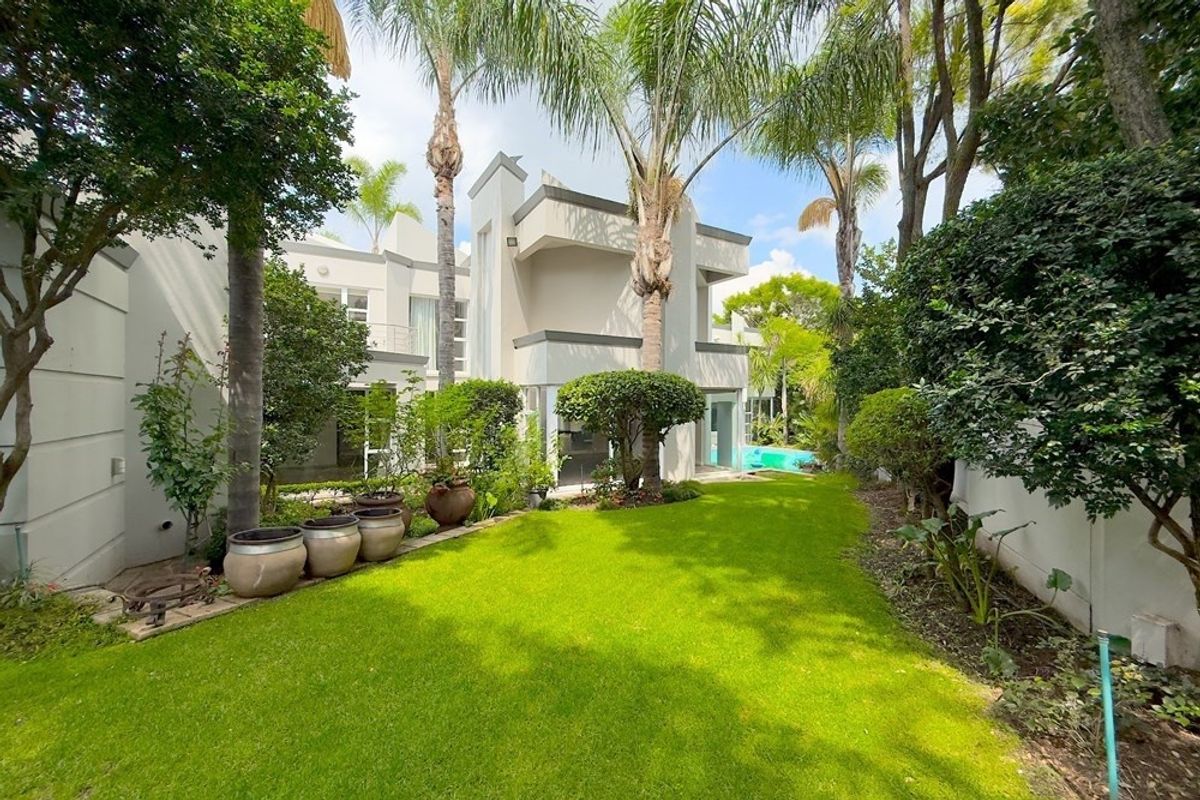 A well-maintained rear garden featuring a lush lawn, mature trees, and a view of the modern multi-story residence with an integrated swimming pool.