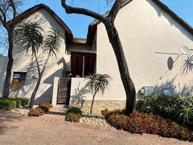 Front elevation of a house featuring a distinctive thatched roof, stone cladding accents, and a paved driveway with established garden landscaping.