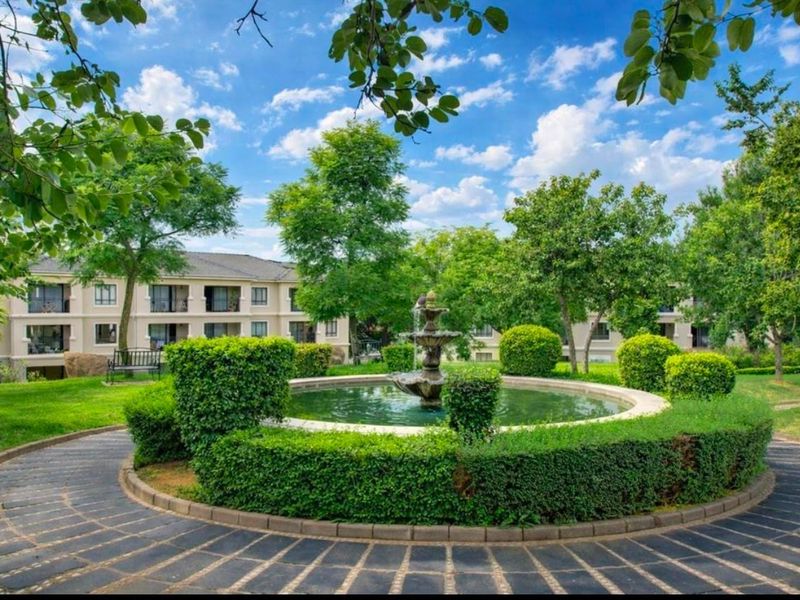 A well-maintained communal garden area featuring a central water fountain, manicured hedges, and paved pathways surrounded by mature trees and apartment building views.
