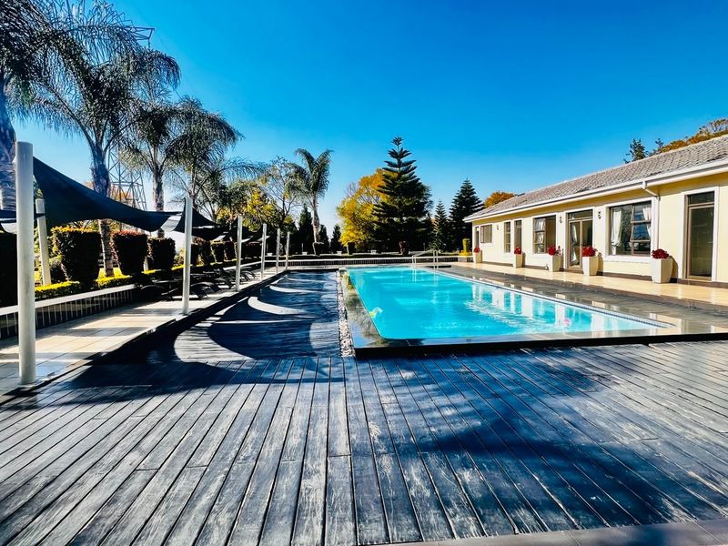 A well-maintained outdoor pool area featuring a rectangular swimming pool, extensive timber decking, and shade sails, adjacent to the apartment building.
