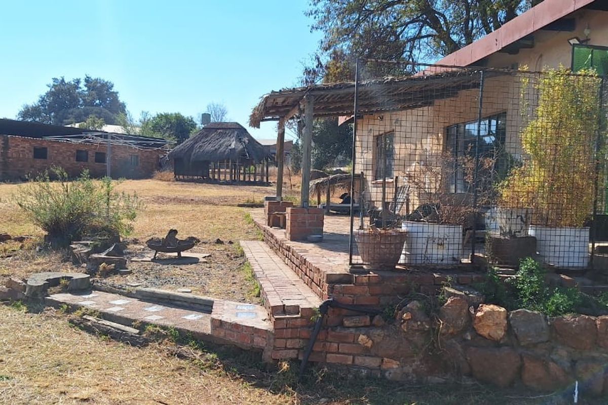 Rear view of the farm property featuring a brick patio, a thatched veranda, and a separate thatched lapa structure set in a rural landscape.