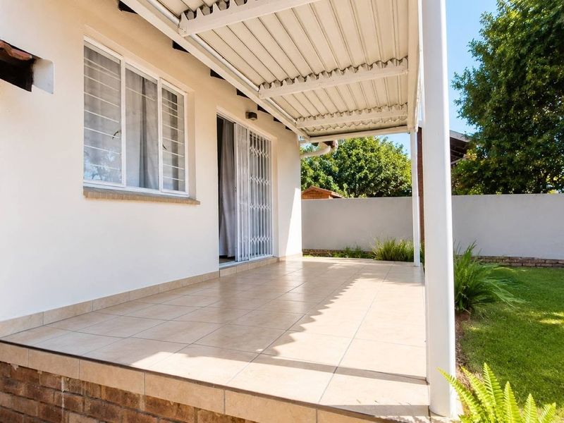 A covered outdoor patio featuring tiled flooring, a metal roof structure, and direct access to the garden through a sliding security gate.