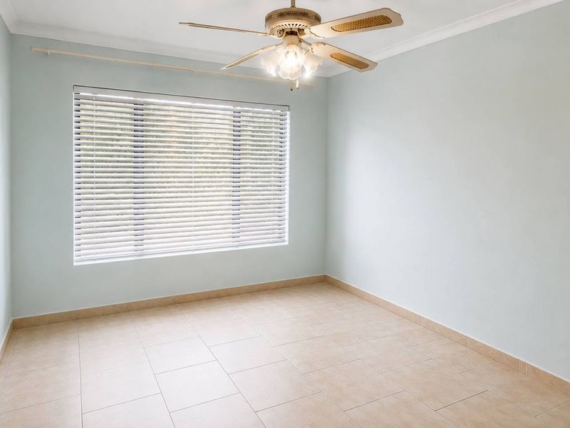 A bright, empty bedroom featuring neutral tiled flooring, a ceiling fan, and venetian blinds, offering a clean canvas for personalization.