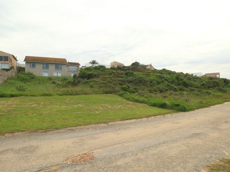 A view of a vacant residential plot featuring sloped terrain and natural coastal vegetation, situated along an unpaved road.