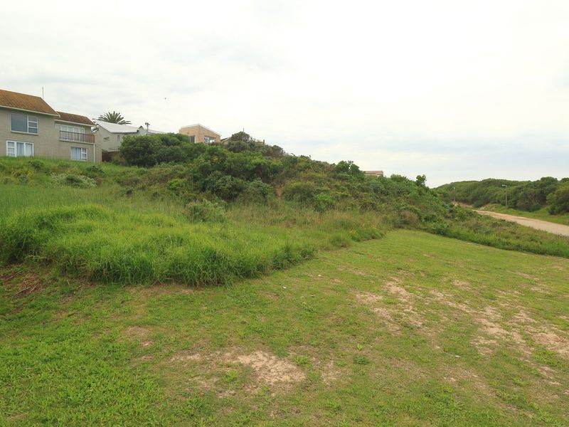 A vacant residential plot featuring sloped terrain, natural vegetation, and direct access to an unpaved road in a coastal neighborhood setting.