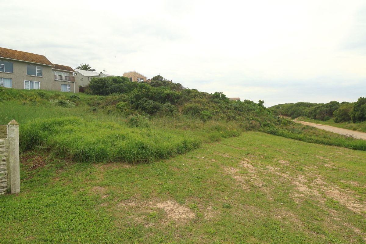 A vacant residential plot featuring sloped terrain, natural vegetation, and direct access to an unpaved road in a coastal neighborhood setting.