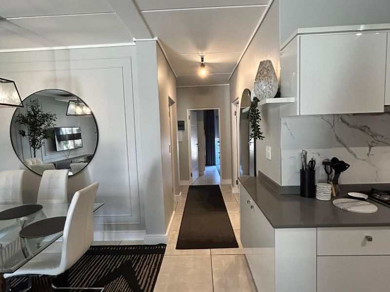 Modern open-plan living and kitchen area featuring sleek cabinetry, stone countertops, and neutral tiled flooring, leading into a central hallway.