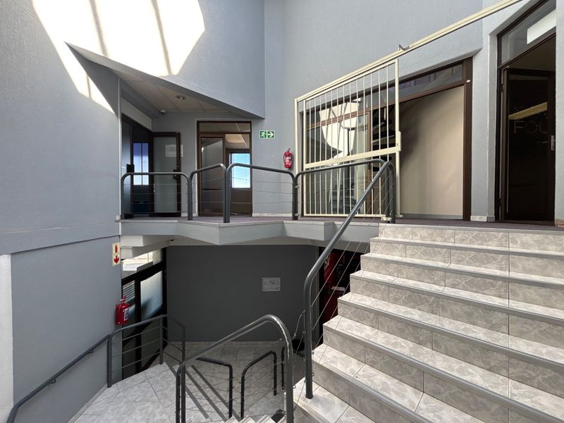 A multi-level office building hallway featuring tiled staircases, metal safety railings, and secure access points with emergency fire safety equipment.