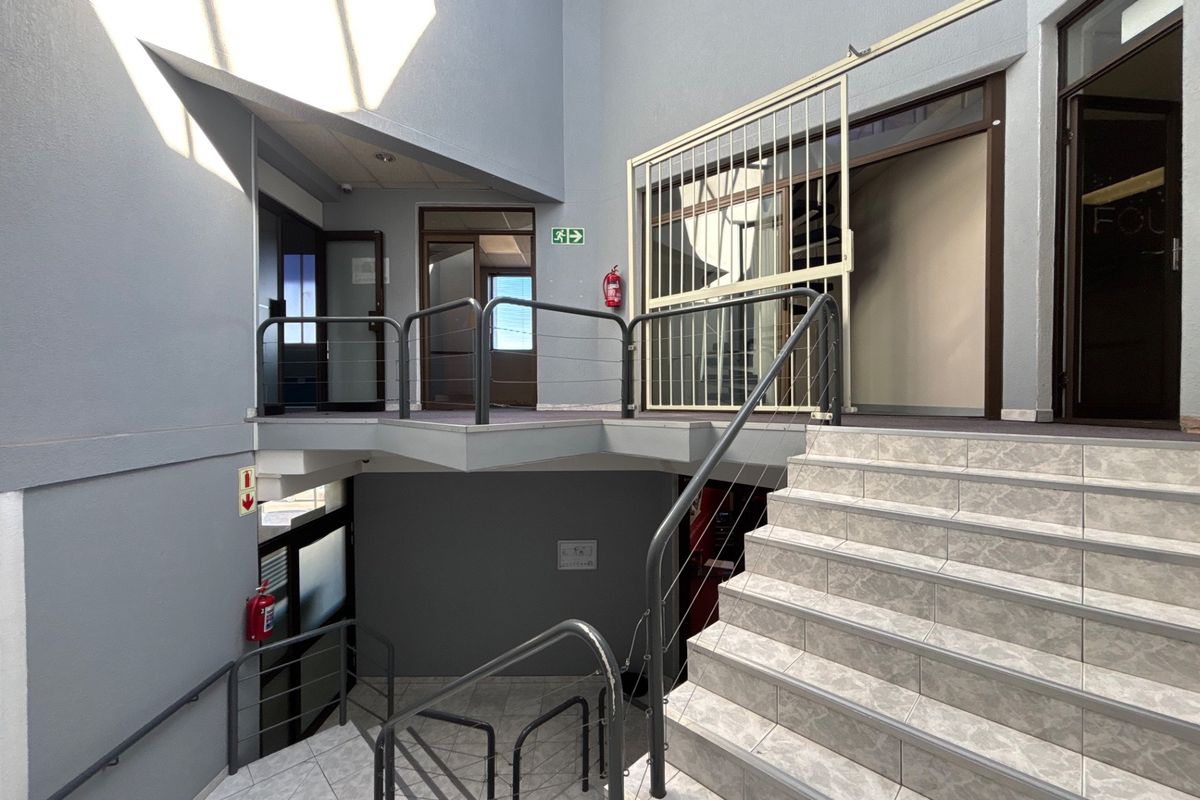 A multi-level office building hallway featuring tiled staircases, metal safety railings, and secure access points with emergency fire safety equipment.