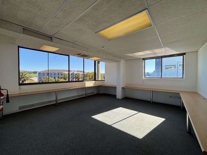 A bright, empty office space featuring a wrap-around built-in desk, large windows with natural light, and a suspended ceiling with recessed lighting.