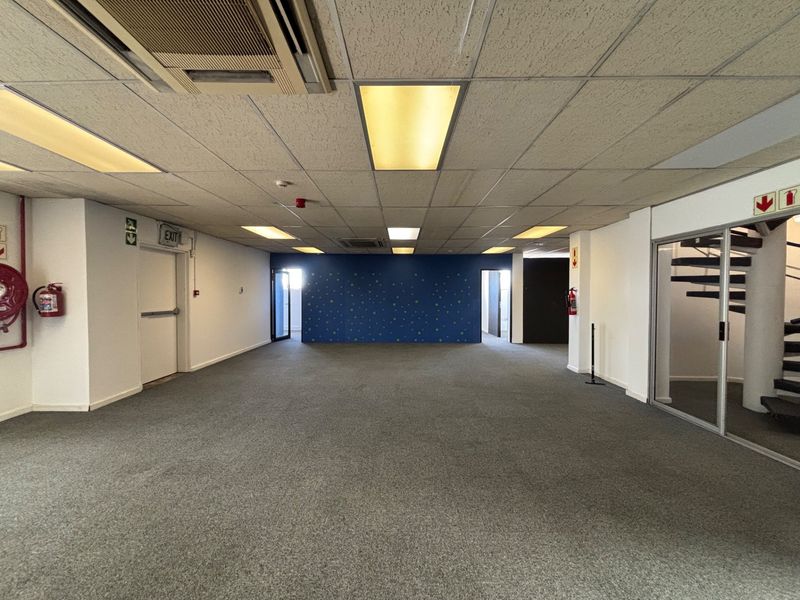 Spacious open-plan office area featuring a suspended ceiling, recessed lighting, carpeted flooring, and an integrated spiral staircase with glass partitioning.