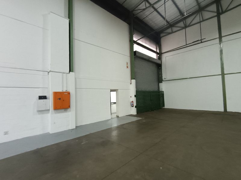 Spacious industrial warehouse unit featuring high ceilings with exposed steel trusses, concrete flooring, and a large roller shutter door for easy access.