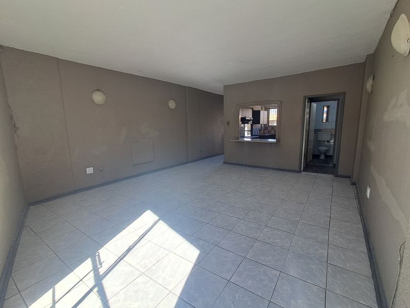 An empty living area featuring tiled flooring, wall-mounted lighting, and a serving hatch connecting to the kitchen, with an adjacent bathroom doorway.