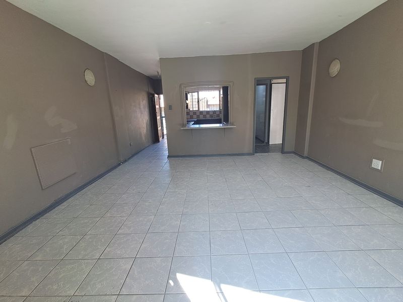 An empty open-plan living area featuring tiled flooring, wall-mounted lighting, and a convenient pass-through window connecting to the kitchen space.