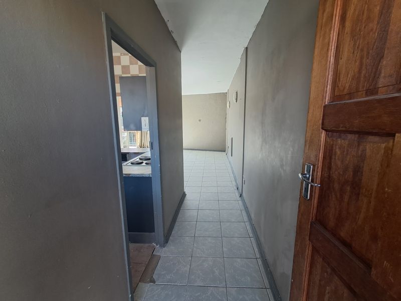 A vacant hallway with tiled flooring leading towards an open plan living area and kitchen, featuring a wooden entrance door.