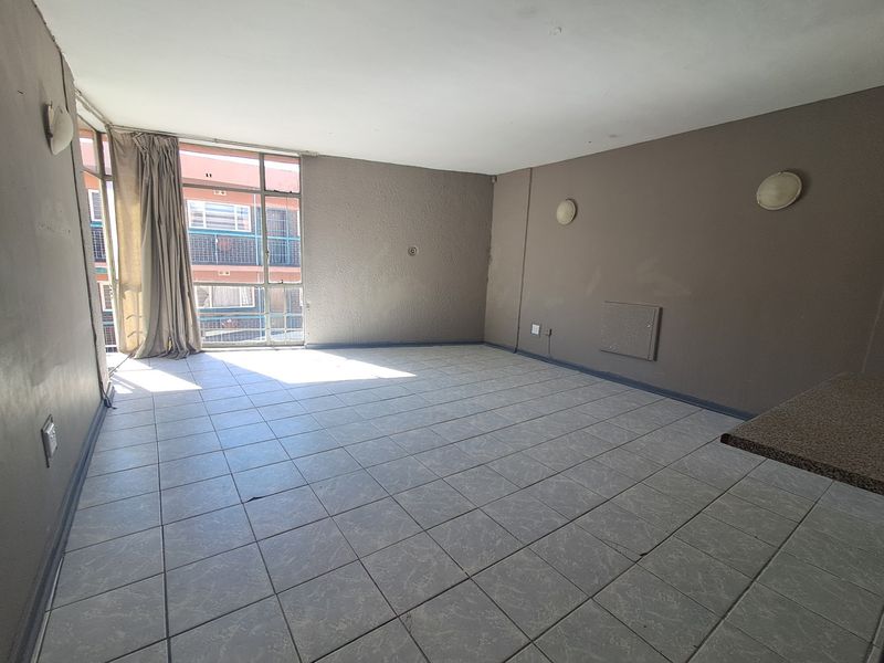 An empty, open-plan living area featuring tiled flooring, wall-mounted lighting, and large windows providing natural light.
