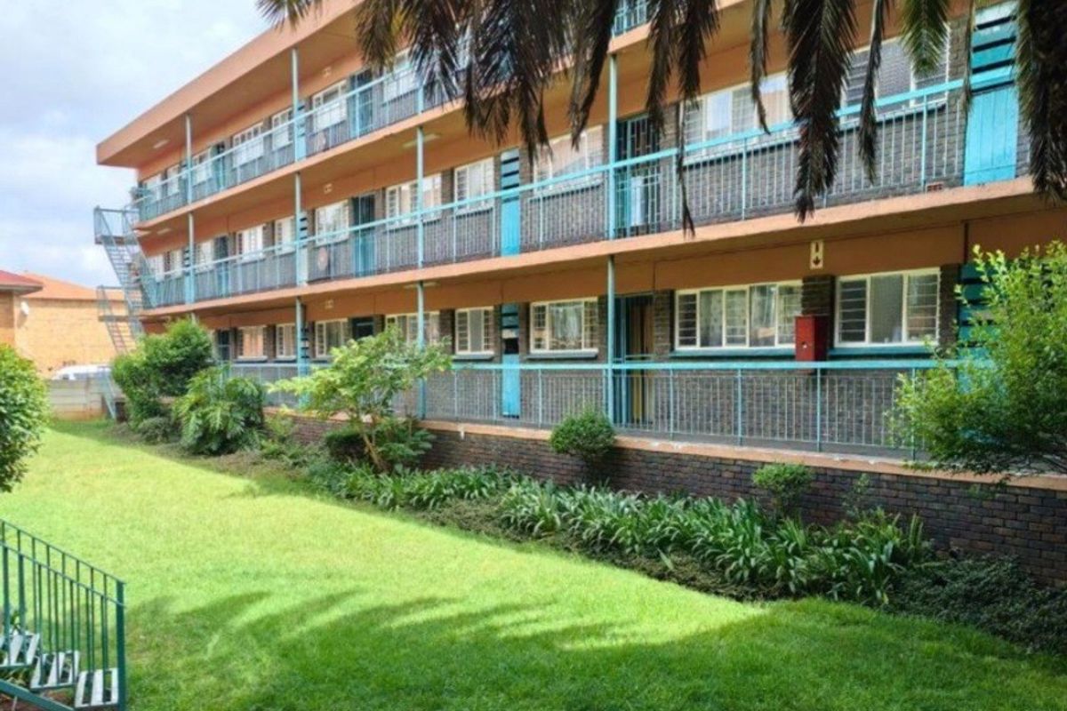 A multi-story apartment building exterior featuring brick facade, metal balcony railings, and a well-maintained communal garden area.