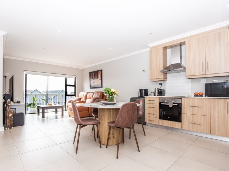 An open-plan living and kitchen area featuring light wood cabinetry, modern built-in appliances, tiled flooring, and a sliding door leading to a balcony.