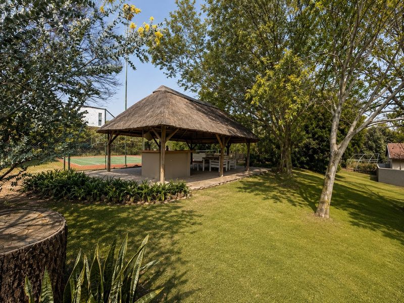 A spacious outdoor thatched lapa featuring a built-in bar area, set within a landscaped garden adjacent to a private tennis court.