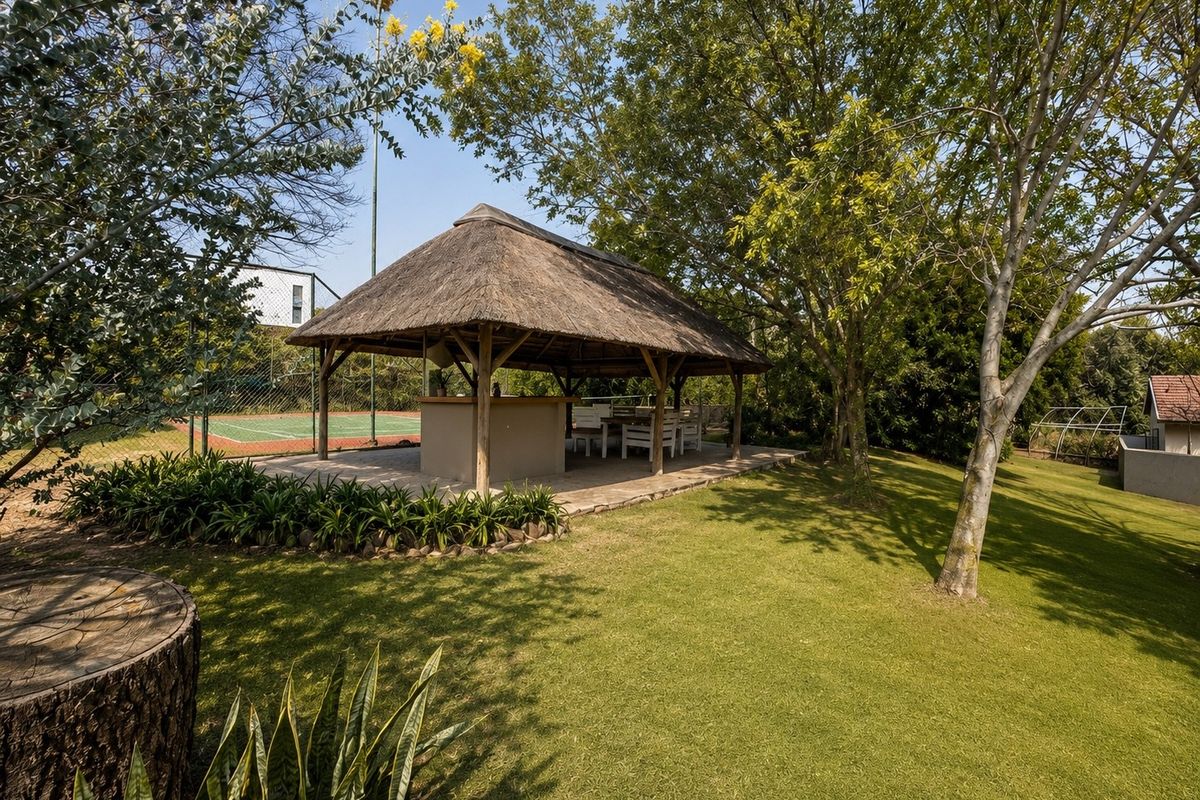 A spacious outdoor thatched lapa featuring a built-in bar area, set within a landscaped garden adjacent to a private tennis court.