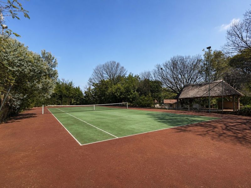 A well-maintained tennis court featuring a green surface with a surrounding red clay area and an adjacent thatched lapa for shade.