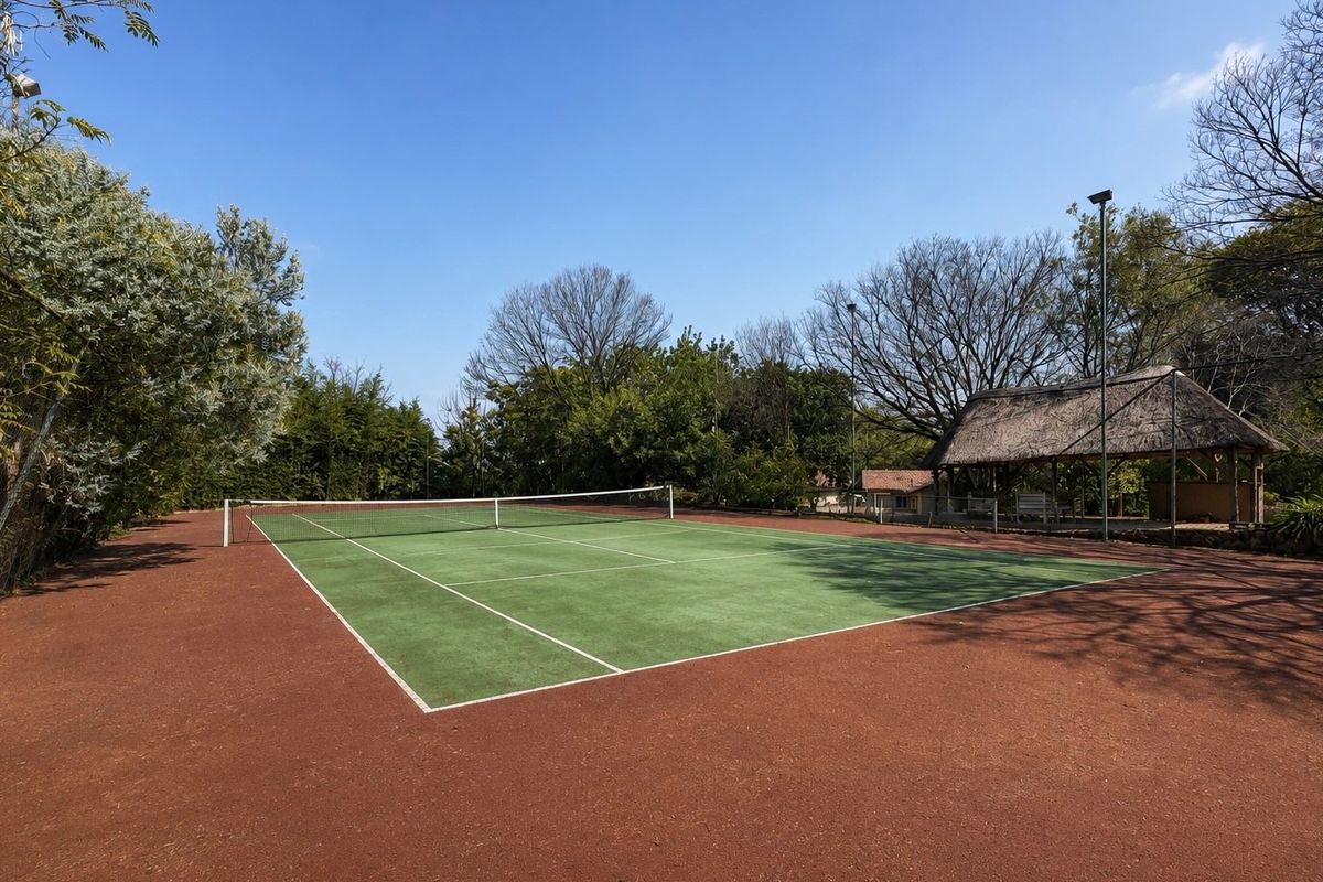 A well-maintained tennis court featuring a green surface with a surrounding red clay area and an adjacent thatched lapa for shade.