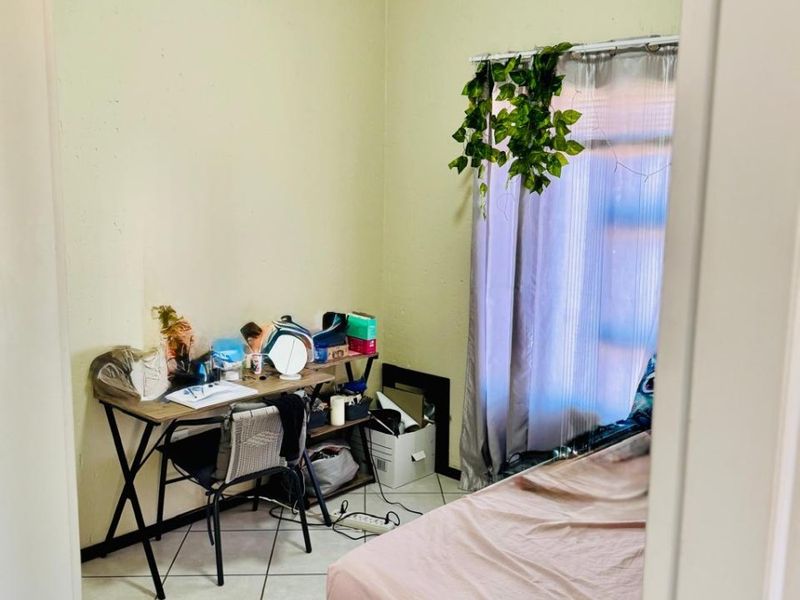 A compact bedroom featuring tiled flooring and a window with a curtain rod, currently used as a combined sleeping and workspace area.