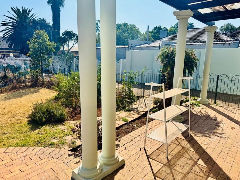 A covered outdoor patio area featuring brick paving and structural columns, overlooking a private garden space.
