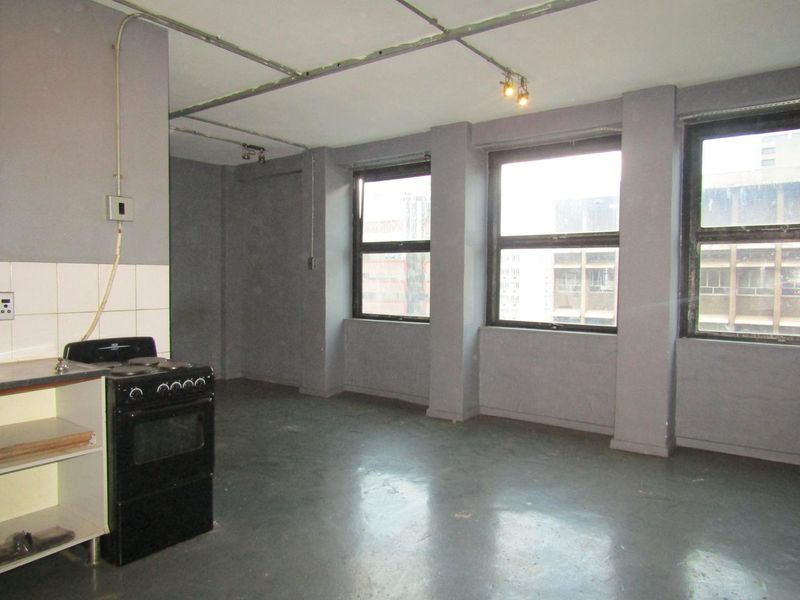 Open-plan living area featuring industrial-style exposed conduit, track lighting, and a basic kitchen corner with a freestanding stove and tiled splashback.