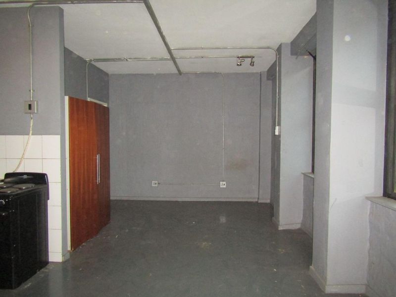 An empty, industrial-style kitchen area featuring exposed electrical conduits, a built-in cupboard, and a basic stove, requiring cosmetic updates.