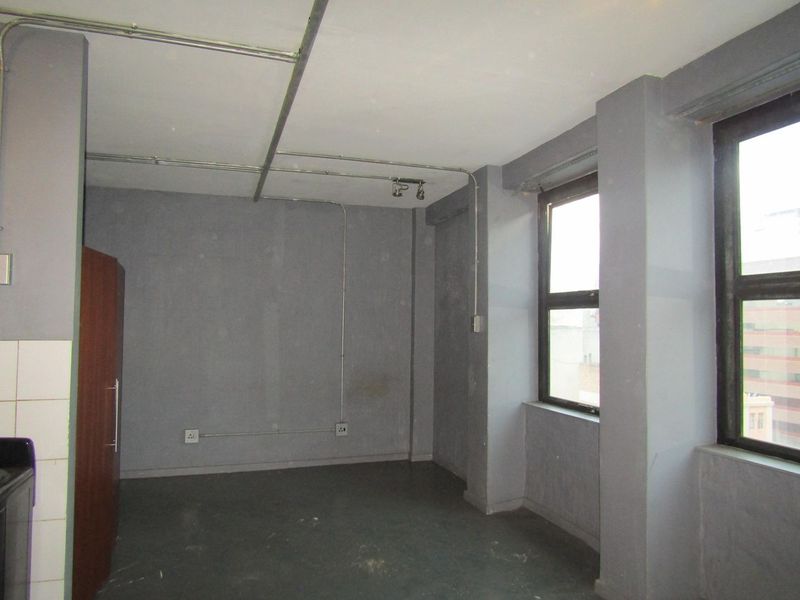 An empty open-plan living area featuring industrial-style exposed conduit, large windows, and neutral grey walls, offering a blank canvas for renovation.