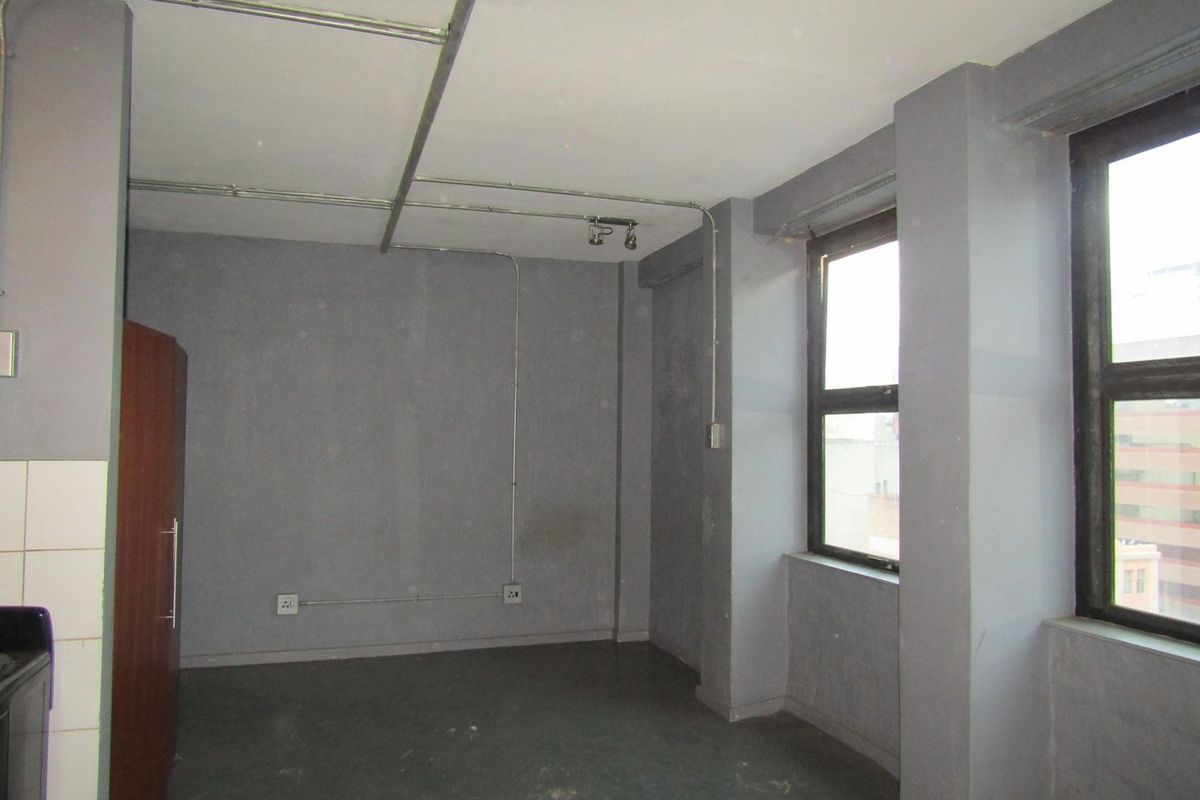 An empty open-plan living area featuring industrial-style exposed conduit, large windows, and neutral grey walls, offering a blank canvas for renovation.