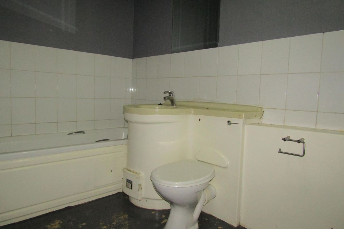 A dated bathroom featuring an integrated vanity and toilet unit with a standard bathtub and tiled splashback, requiring modernization.