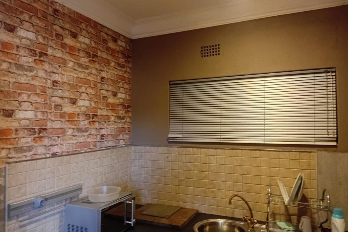 A functional kitchen area featuring a stainless steel sink, tiled splashback, and decorative brick-effect wall finish with venetian blinds.