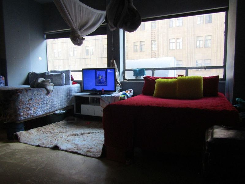 A bedroom featuring large windows with roller blinds, offering natural light and urban views. The space requires cosmetic updates to improve its overall condition.