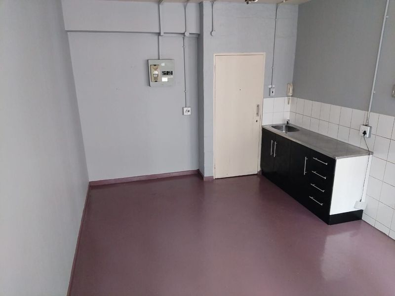 A compact, empty kitchen area featuring basic cabinetry, a stainless steel sink, tiled splashback, and exposed surface-mounted electrical conduits.