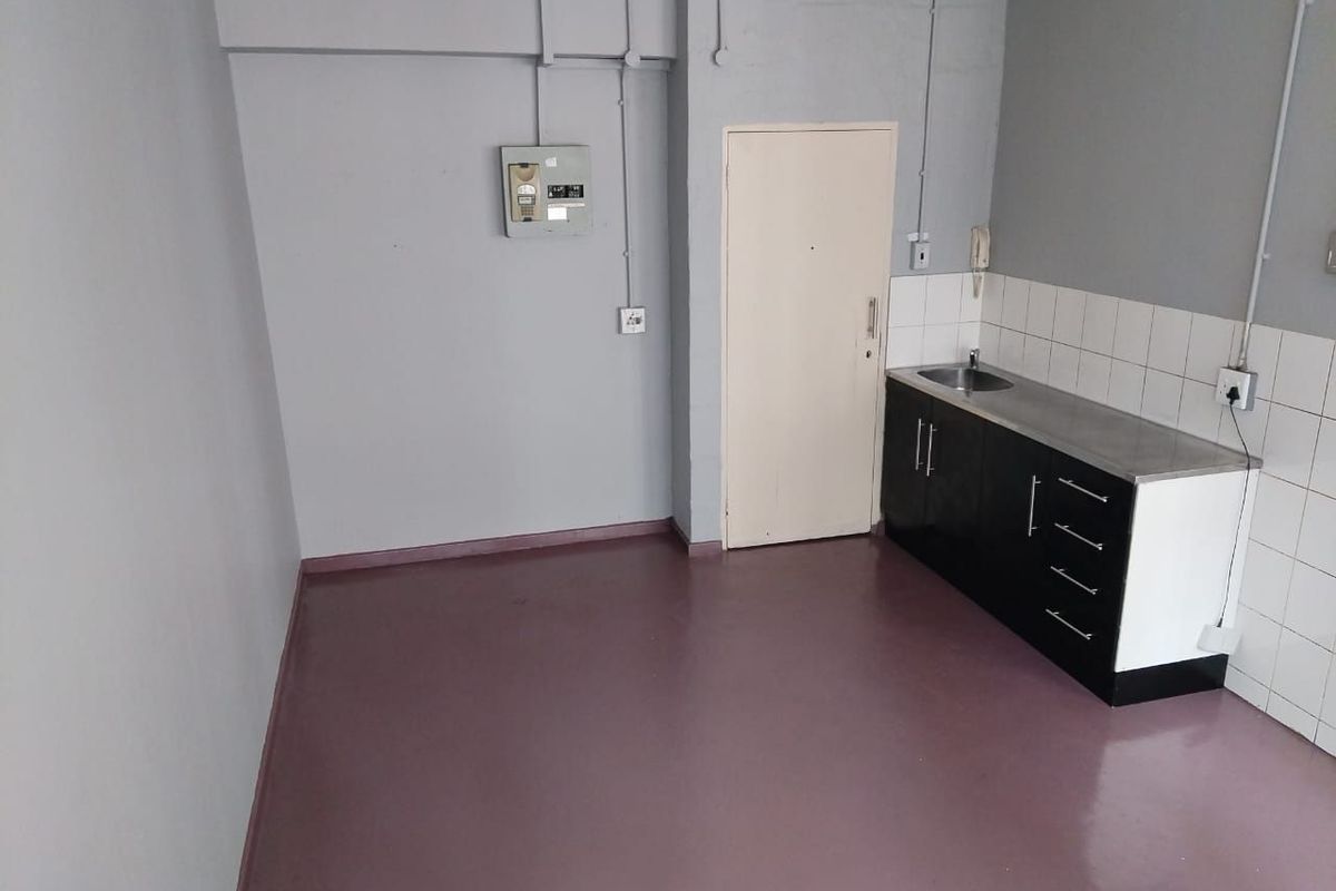 A compact, empty kitchen area featuring basic cabinetry, a stainless steel sink, tiled splashback, and exposed surface-mounted electrical conduits.