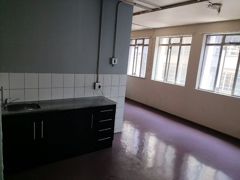 An open-plan living and kitchen area featuring a basic kitchenette with a stainless steel sink, tiled splashback, and large windows providing natural light.